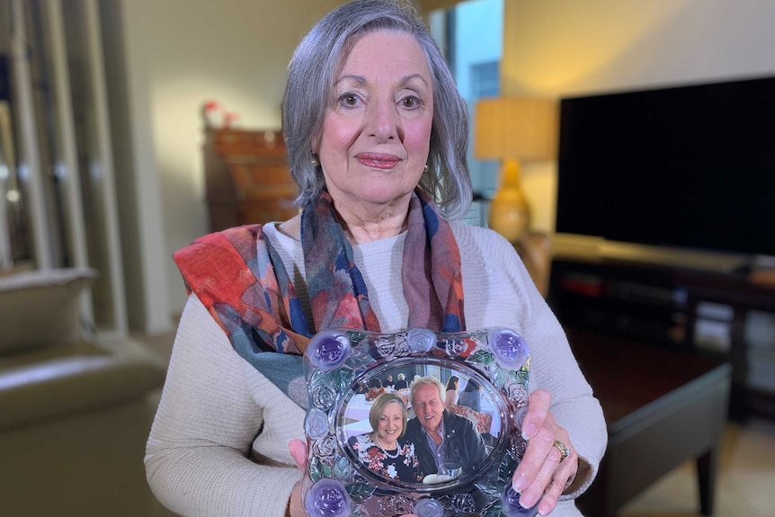 Mandy Whitlock sits looking at the camera from her house, holding a photo of her and her husband together.