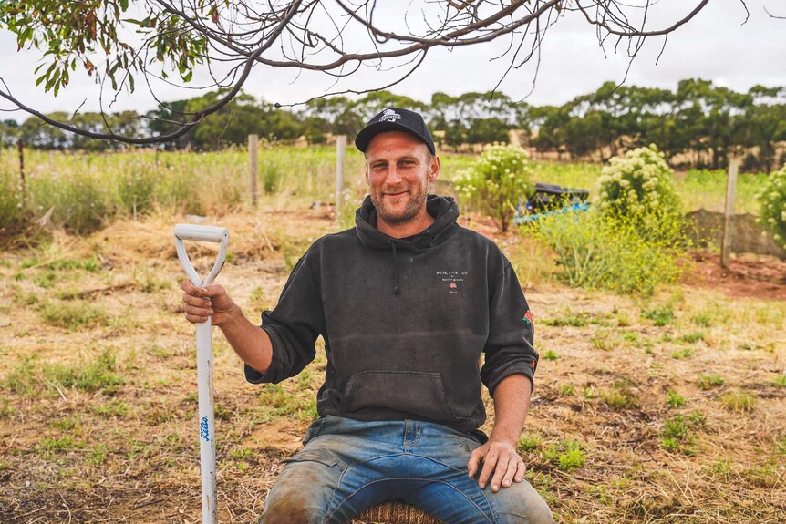 A man wearing a worn hoodie and cap sits on an old wooden chair grinning, holding a shovel in a green garden.