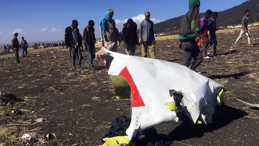 Several people walk past part of a crumpled metal plane part at the scene of a plane crash.