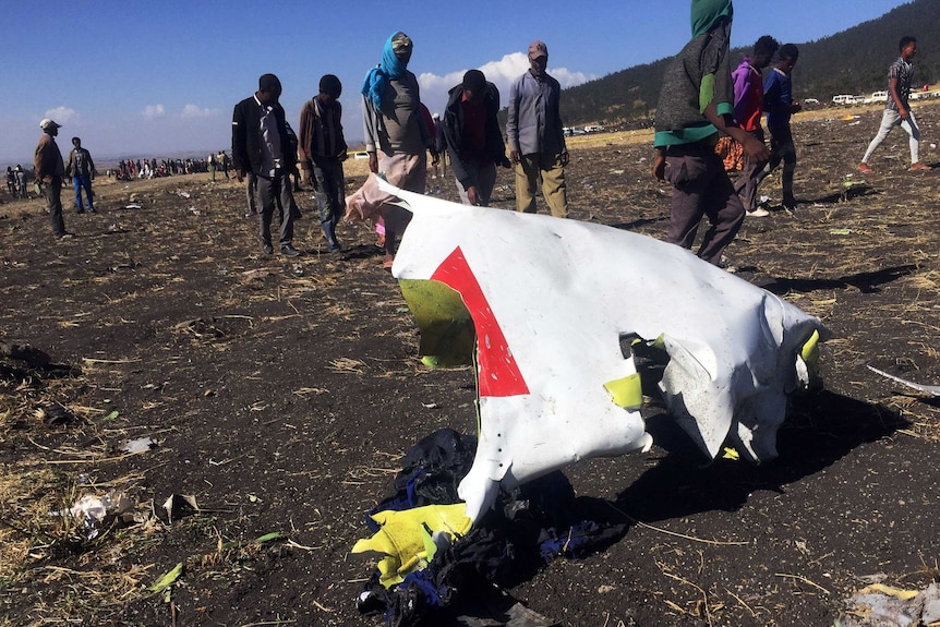 Several people walk past part of a crumpled metal plane part at the scene of a plane crash.