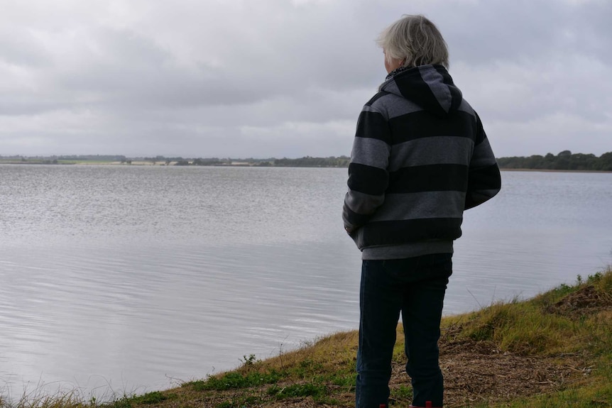 The back of a woman with short grey hair, a stripey jumper and red gumboots in front of a lake.