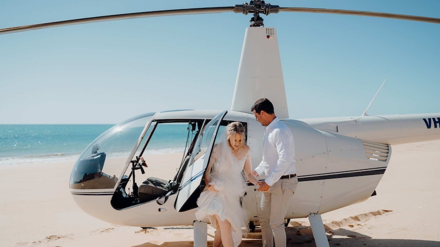 A woman and man exit a helicopter on a beach, dressed for their wedding.