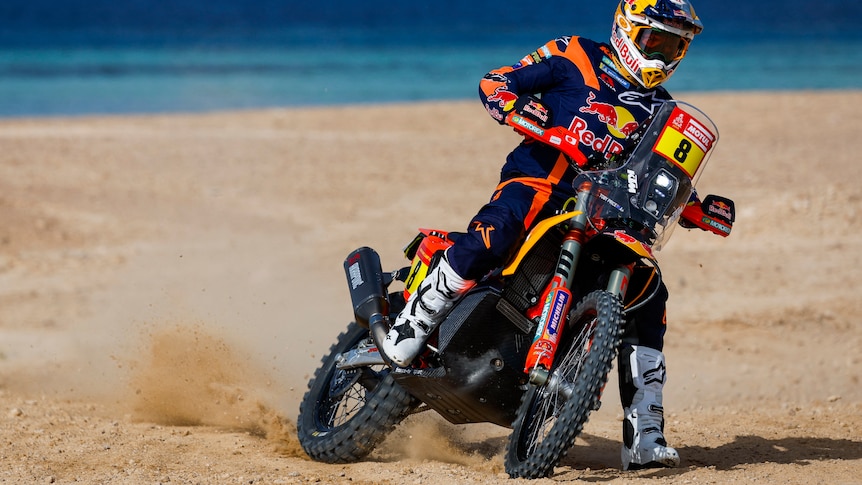 A rally motorbike turning on a beach, with the sea in the background.