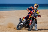 A rally motorbike turning on a beach, with the sea in the background.