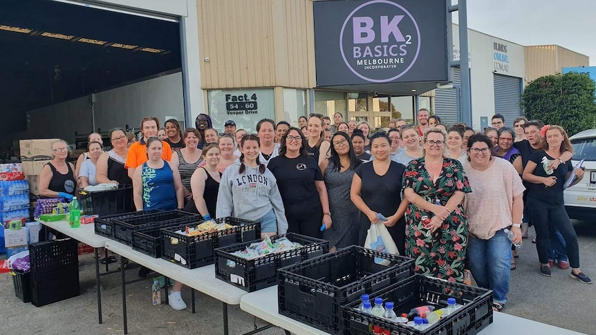 About 50 people, mostly women, stand in a carpark outside a warehouse beside long tables filled with drinks and snacks.