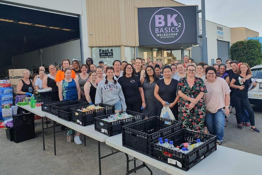 About 50 people, mostly women, stand in a carpark outside a warehouse beside long tables filled with drinks and snacks.