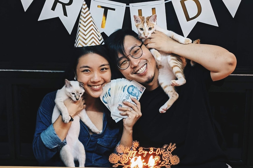 Ken Yip with his wife holding their two cats, wearing birthday hats. 