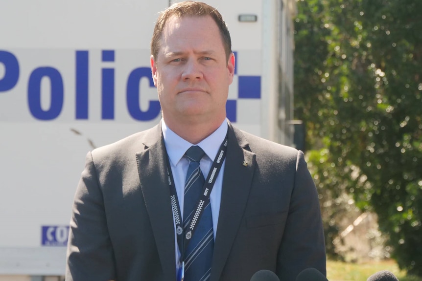 A man in a suit, with a police lanyard on, in front of microphones