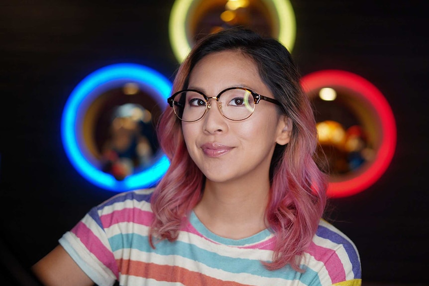 A bespectacled woman with long hair, top half dark, bottom half dyed pink poses in front of wall with three neon circular lights