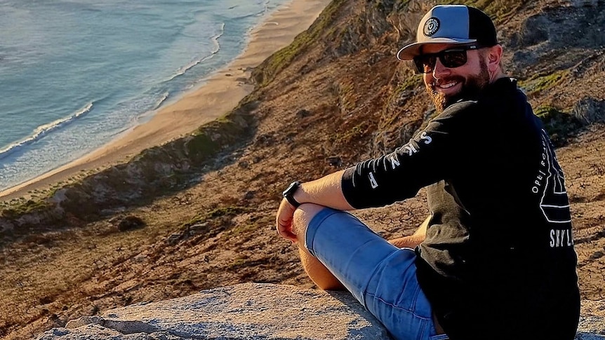 Man sitting on hill by ocean, turning around to look at camera.