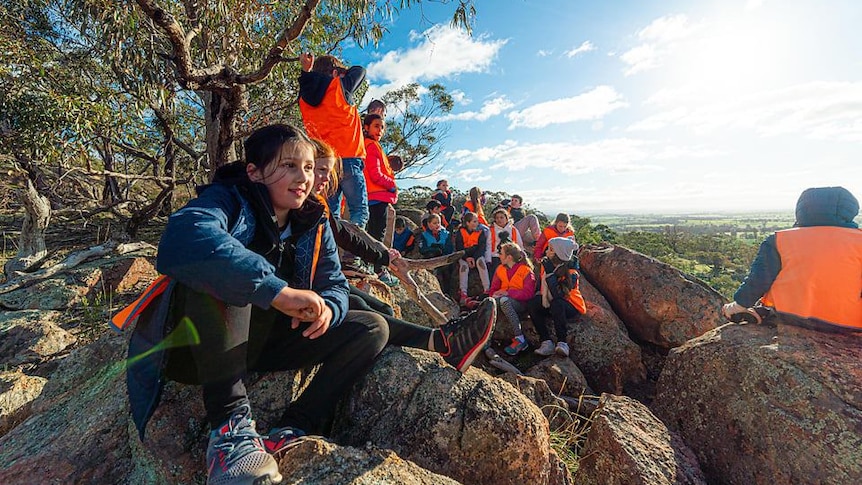 Kids smile as they sit on a rocky montain top with sunny views of green farmland all around, Maldon Victoria.