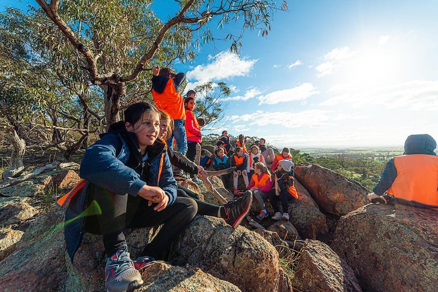 Kids smile as they sit on a rocky montain top with sunny views of green farmland all around, Maldon Victoria.