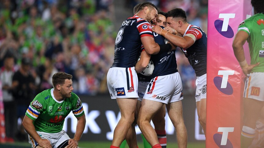The Sydney Roosters players stand and celebrate a try as a Canberra Raiders player kneels down next to them.