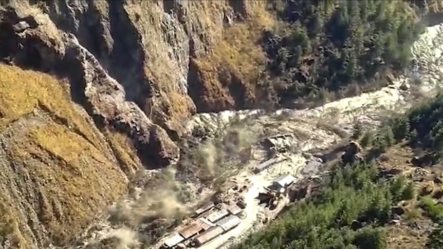 An aerial shot of a mountain valley shows a flooded river surging over houses and trees.