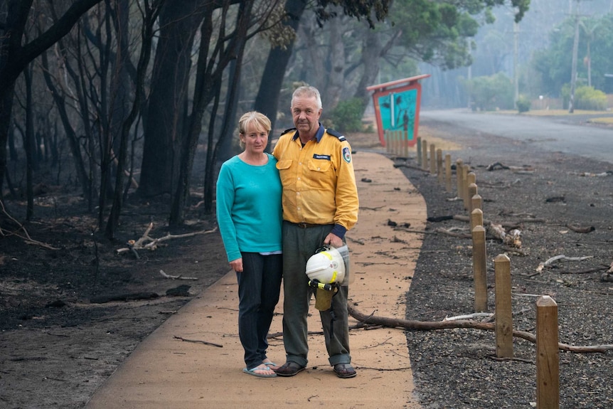 A man in a firefighters uniform and a woman pose with arms around one another on a footpath next to burnt out forest.