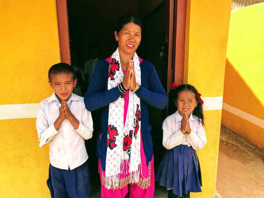 A woman and two children stand with their hands in front of them, in a position to give thanks, in front of a yellow building.