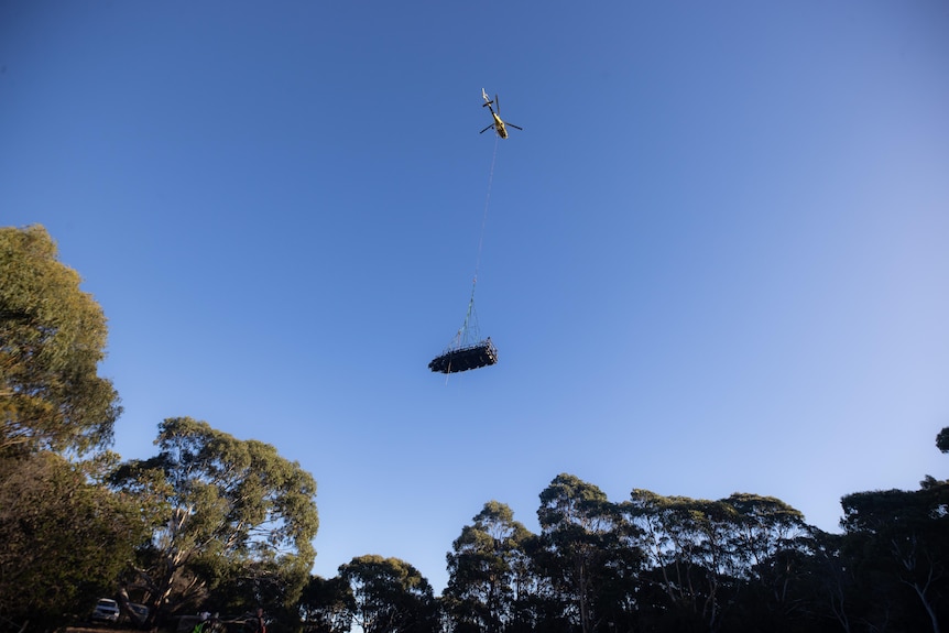 Helicopter with long rope carries boat made from marine debris over trees 