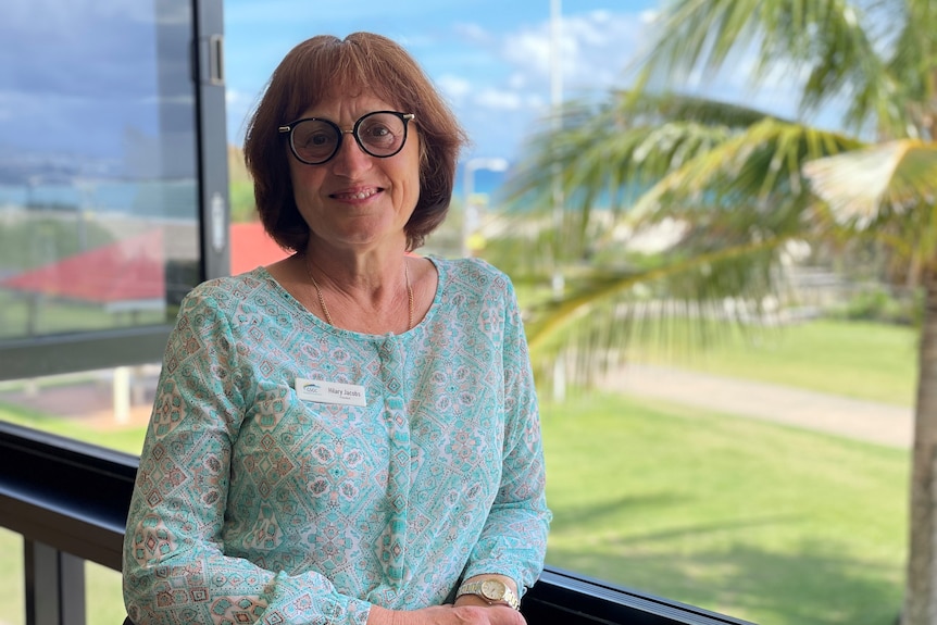 A bespectacled woman of late middle-age stands smiling on a balcony overlooking a lawn.