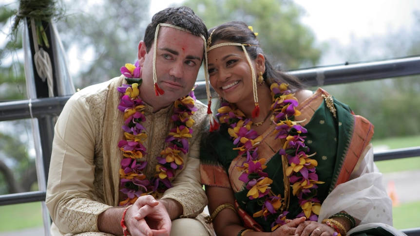 A man and woman pose together on wedding day, wearing garlands of flowers.