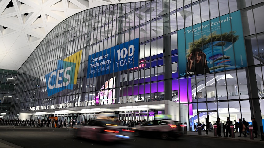 A wide shot of the front of a large exhibition centre with CES branding on it. Cars and people go past the building