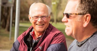 Elderly man with glasses and white balding hair sits next to senior man on park bench