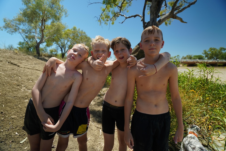 Four boys with their arms around each other stand on the edge of a brown river