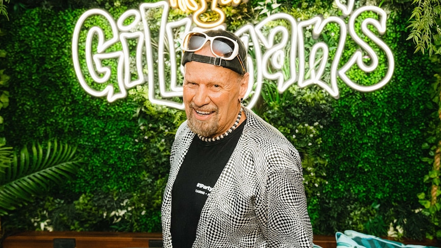 Older man with cap worn backwards and loud patterned shirt in front of Gilligan's sign
