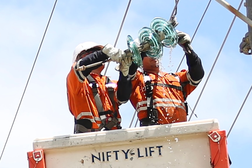 Two power workers in a cherry picker drain water from an insulator on a power pole.