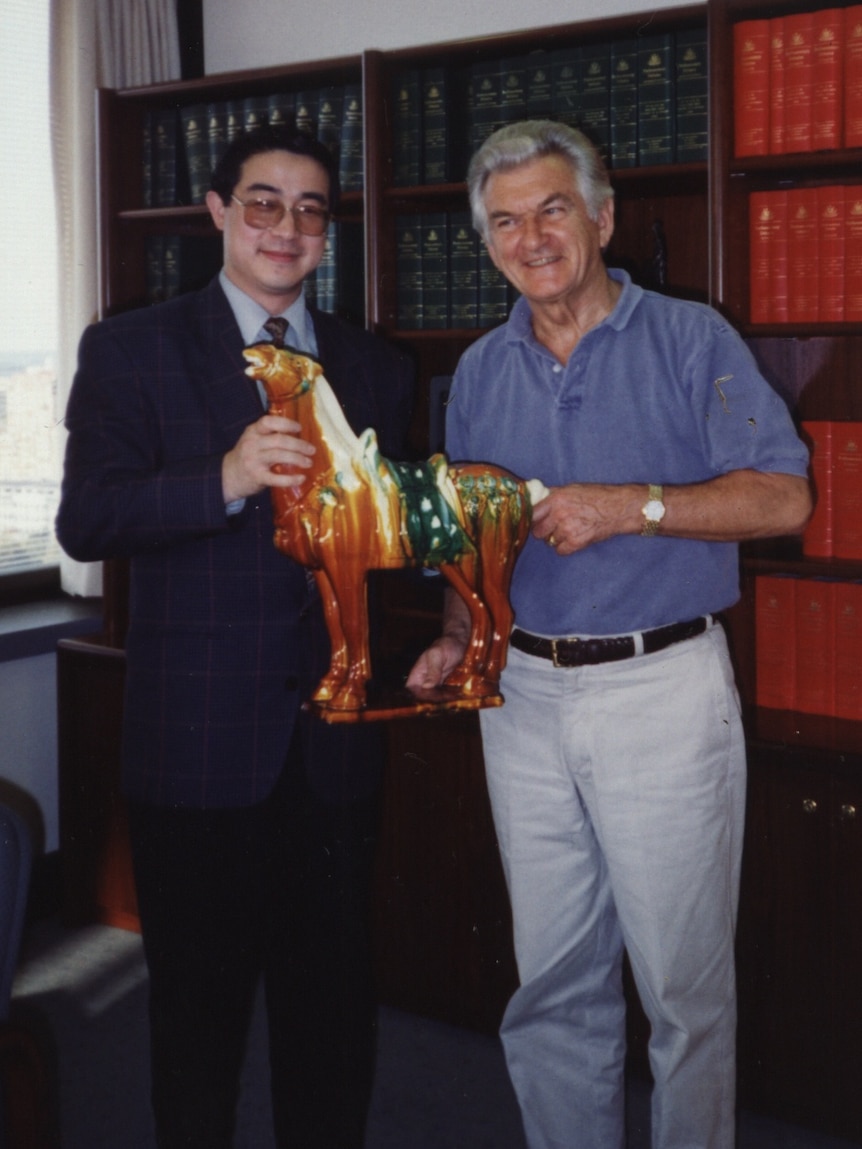 Jun Yang, left, stands next to Bob Hawke, right, in front of a bookshelf in a room. They are holding a small statue of a horse.