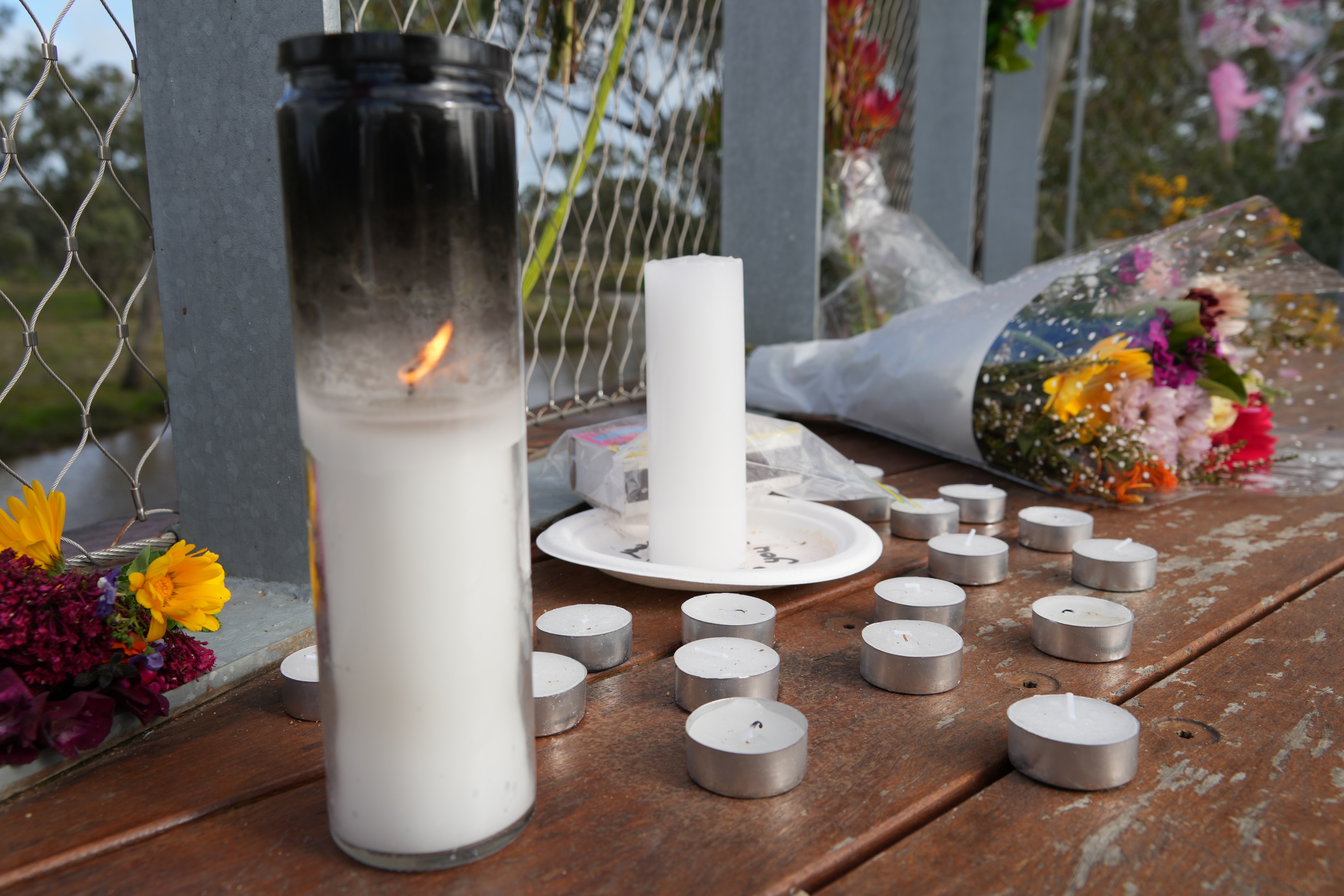 Candles and flowers on a timber deck at a memorial in front of a river.