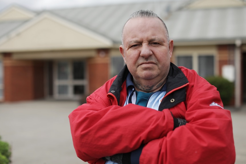 A man wearing a red jacket, frowns and folds his arms as he stands outside a low set brick building.