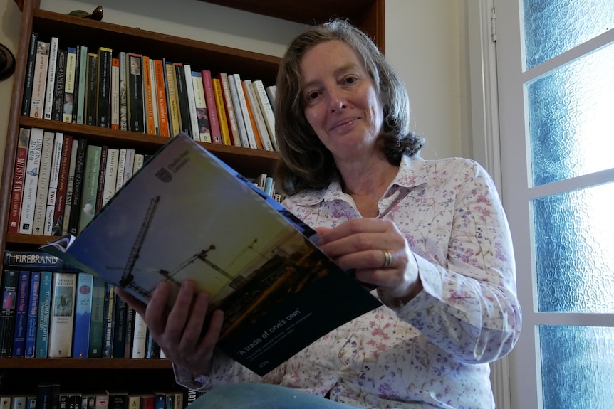 A woman sits on a chair in front of a bookshelf, holding a document titled 'A trade of one's own'