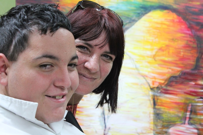 Close up of teen boy and his mum, faces close and smiling, with self-portrait painting behind.