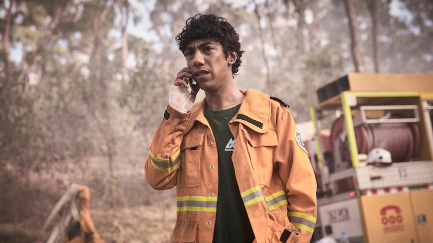 Actor in orange firefighter outfit standing in front of truck talking on a phone.