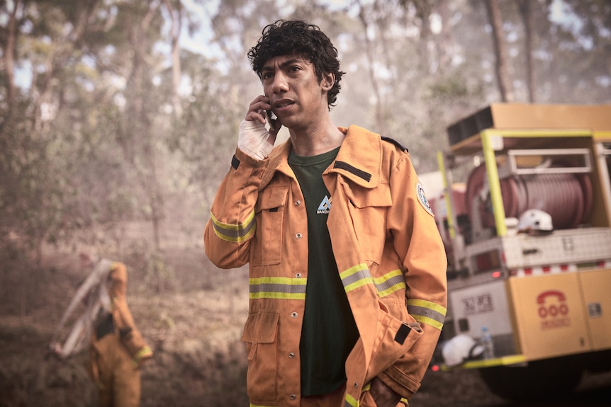Actor in orange firefighter outfit standing in front of truck talking on a phone.