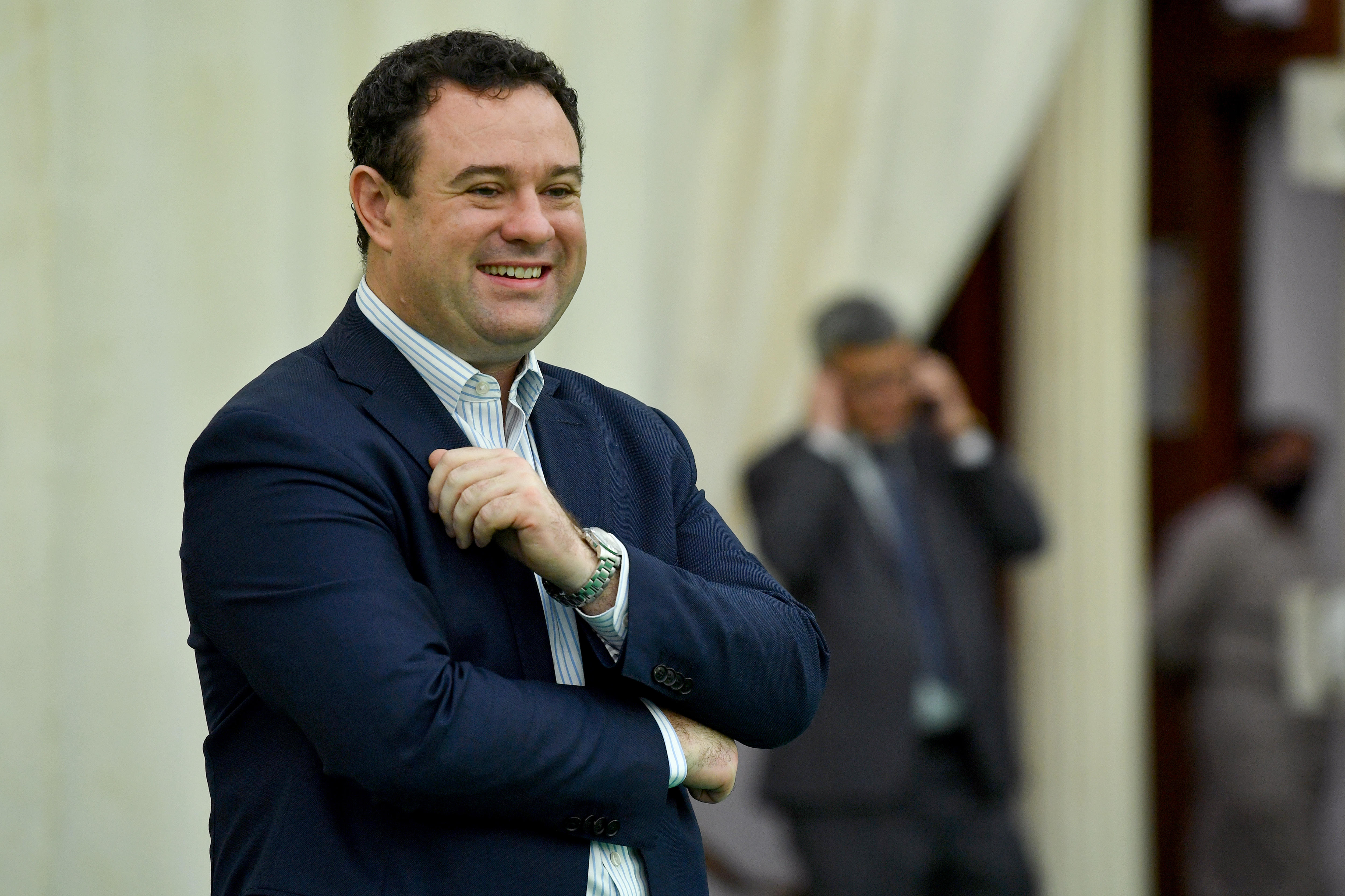 A man wearing a dark blue suit smiles in front of a large fancy white curtain indoors.