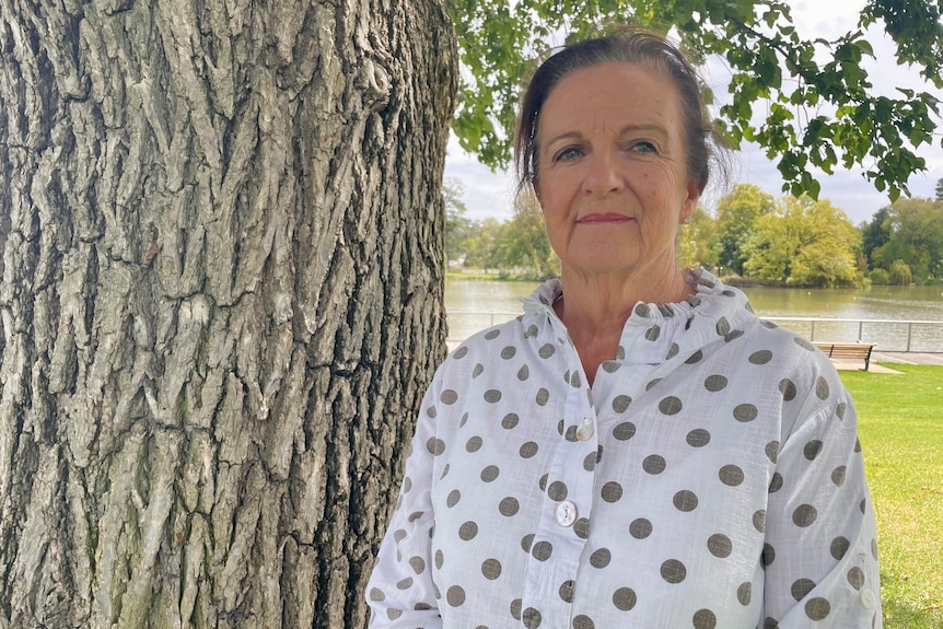 Older woman in white shirt with grey dots standing next to a tree.