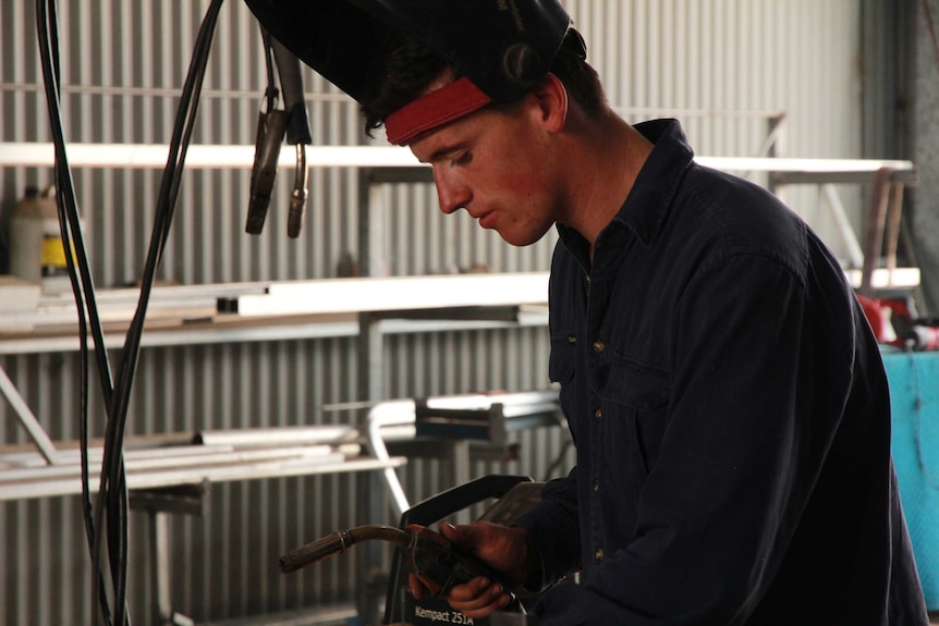 A man leans forward as he prepares to weld a piece of metal in a workshop.
