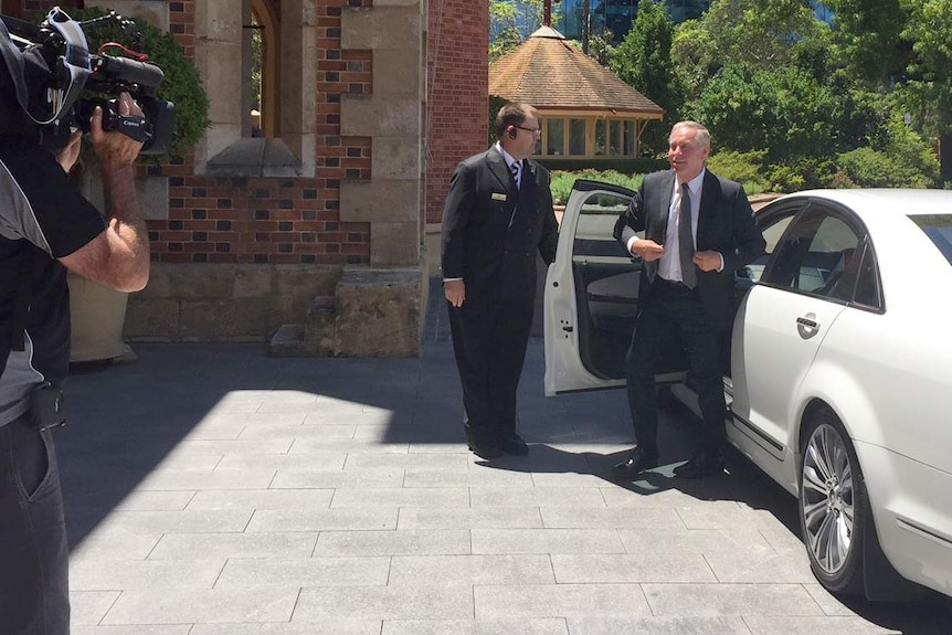 Colin Barnett gets out of a white car on the passenger side with a man holding the door and a cameraman filming.
