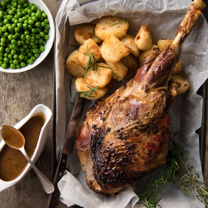 A roasted leg of lamb on a tray with potatoes, a bowl of peas and a jug of gravy.