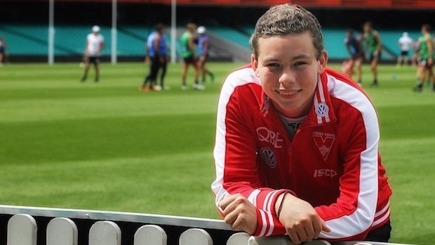 Cooper Rice-Brading, wearing a Swans track suit top, leans on the oval boundary fence while the Swans AFL squad train