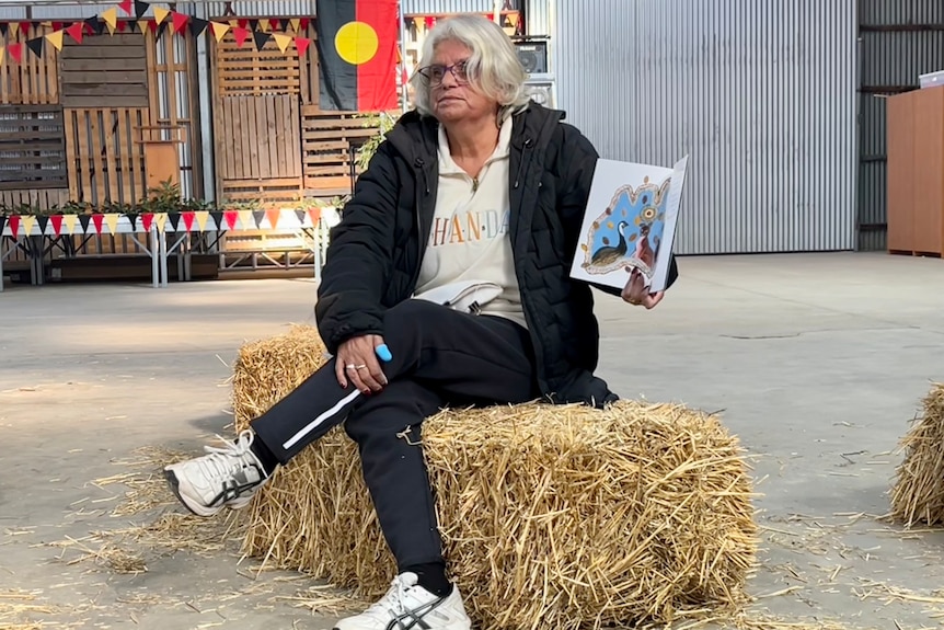 A woman with white hair and glasses and tracksuit sits cross legged on a hay bale holding a book.