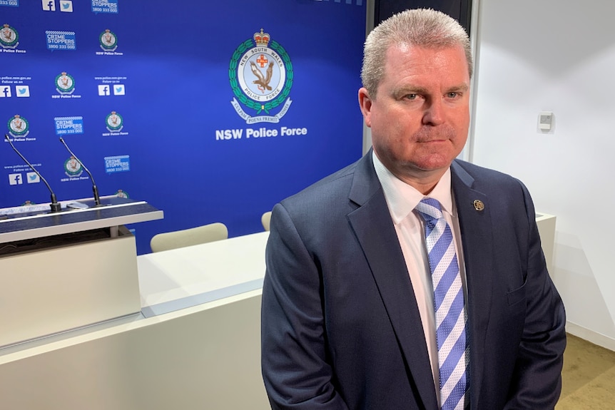 A police officer stands in front of a sign saying 'NSW Police Force'. He is not smiling