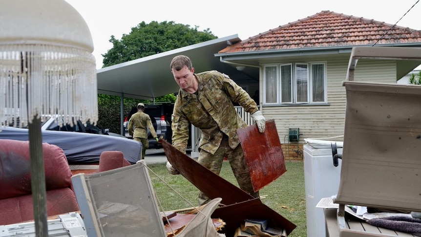 An army officer pics up furniture and puts it on the lawn in front of a house. 