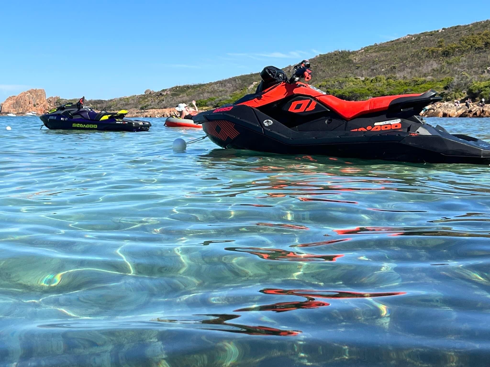 Two jet-skis are in pristine clear blue water, in ocean, with coastline in the background