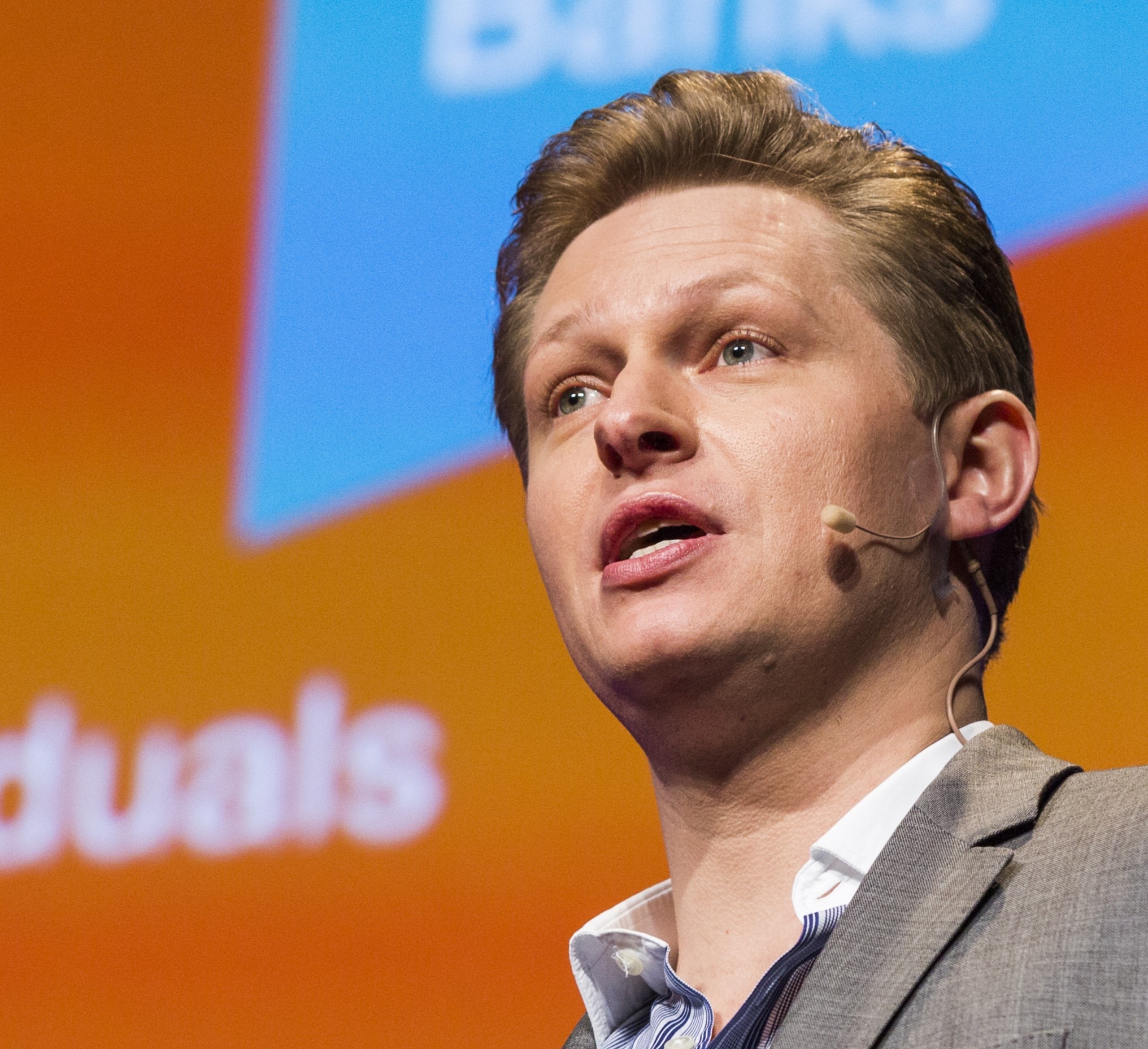 Man with microphone on and suit looks past the camera like he is talking to an audience