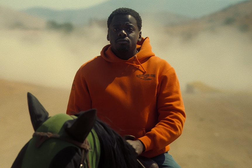 A young man wearing an orange jumper rides a horse in a dusty landscape