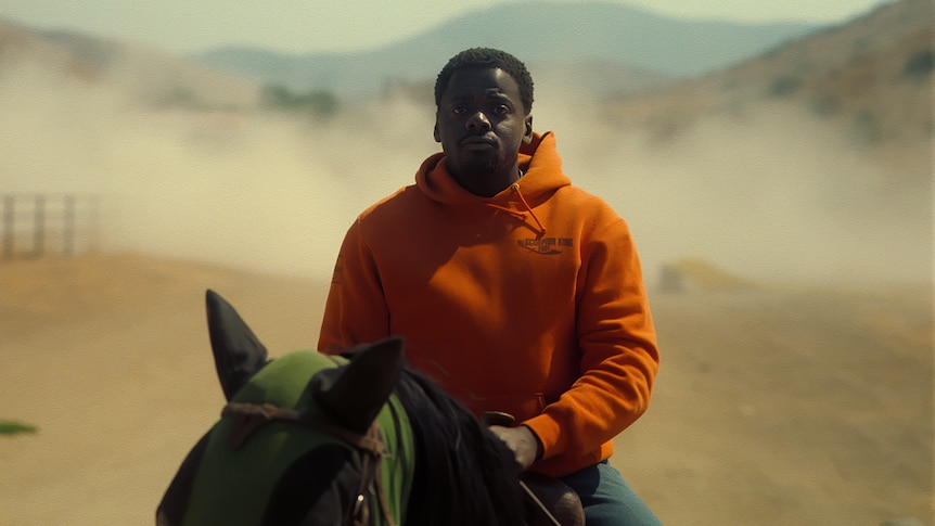 A young man wearing an orange jumper rides a horse in a dusty landscape