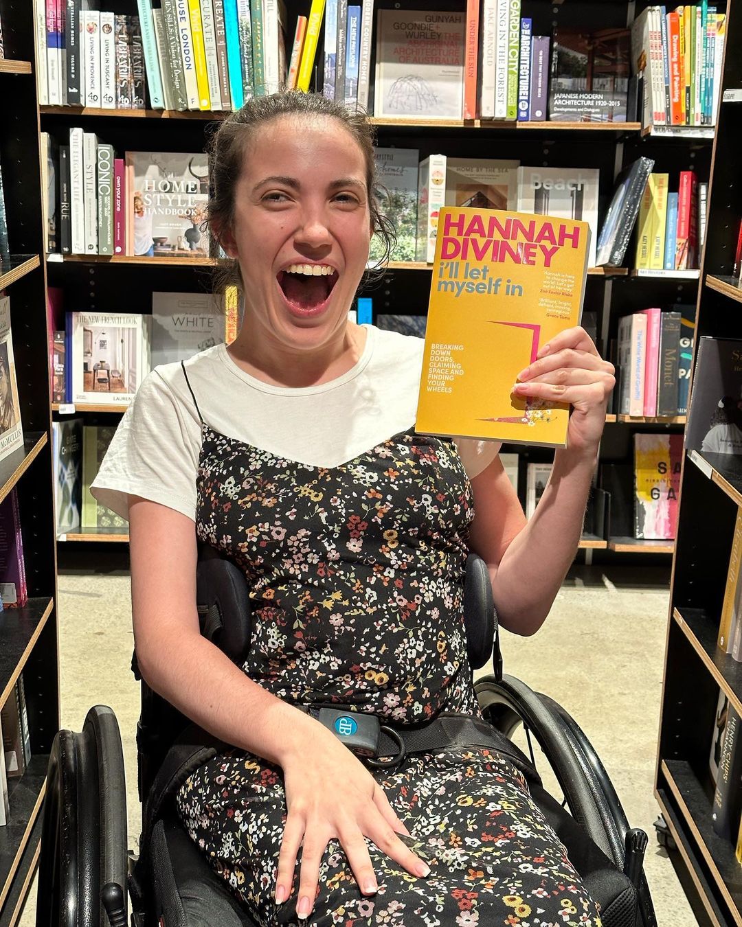 Hannah Diviney in a bookshop smiling as she holds a copy of her book 'I'll Let Myself In'. She is sitting in a wheelchair. 
