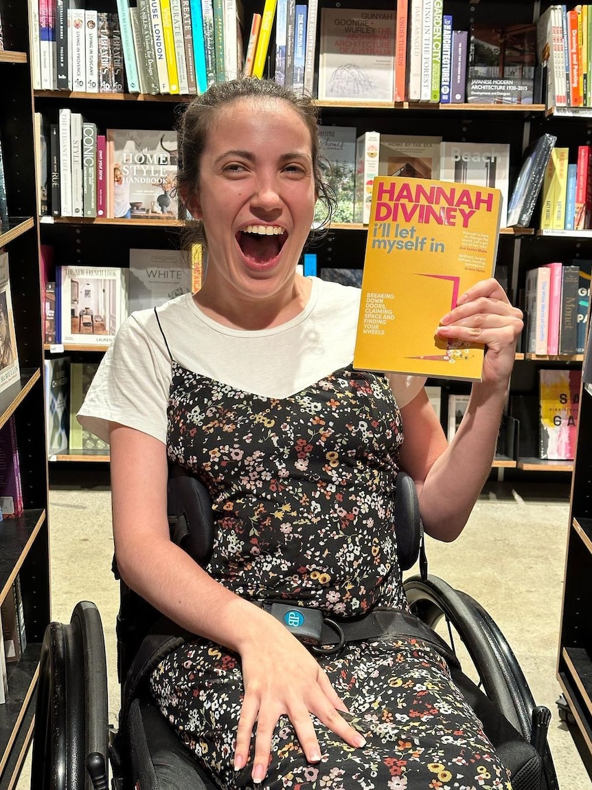 Hannah Diviney in a bookshop smiling as she holds a copy of her book 'I'll Let Myself In'. She is sitting in a wheelchair.